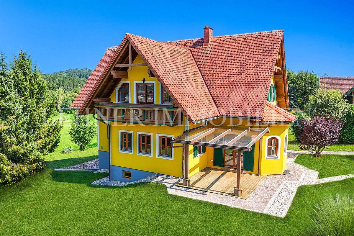 Charmantes Landhaus mit gelber Fassade, Ziegeldach und überdachter Holzterrasse, umgeben von grüner Natur.