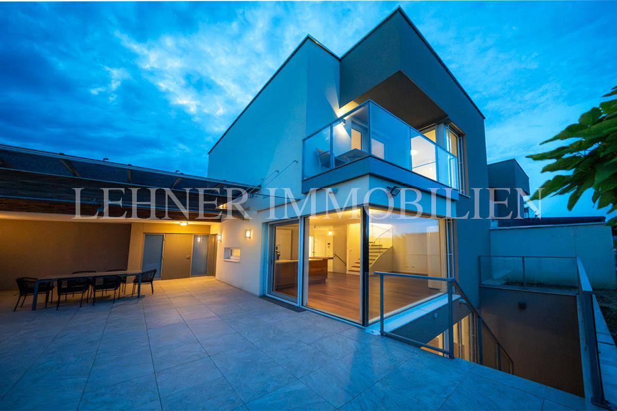 Moderne Hausfassade bei Nacht mit beleuchtetem Balkon und großen Glasflächen, die den Innenraum zeigen.