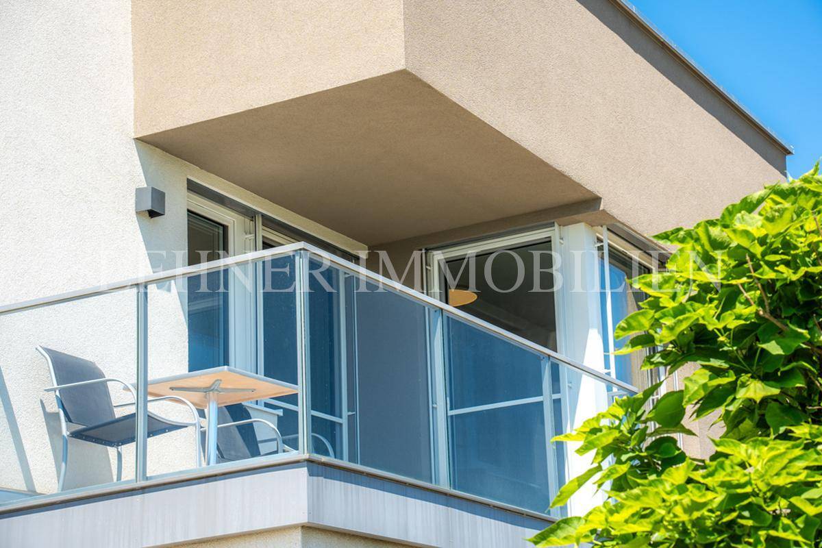 Moderner Balkon mit Glasgeländer und Sitzgelegenheiten, umgeben von üppigem Grün.