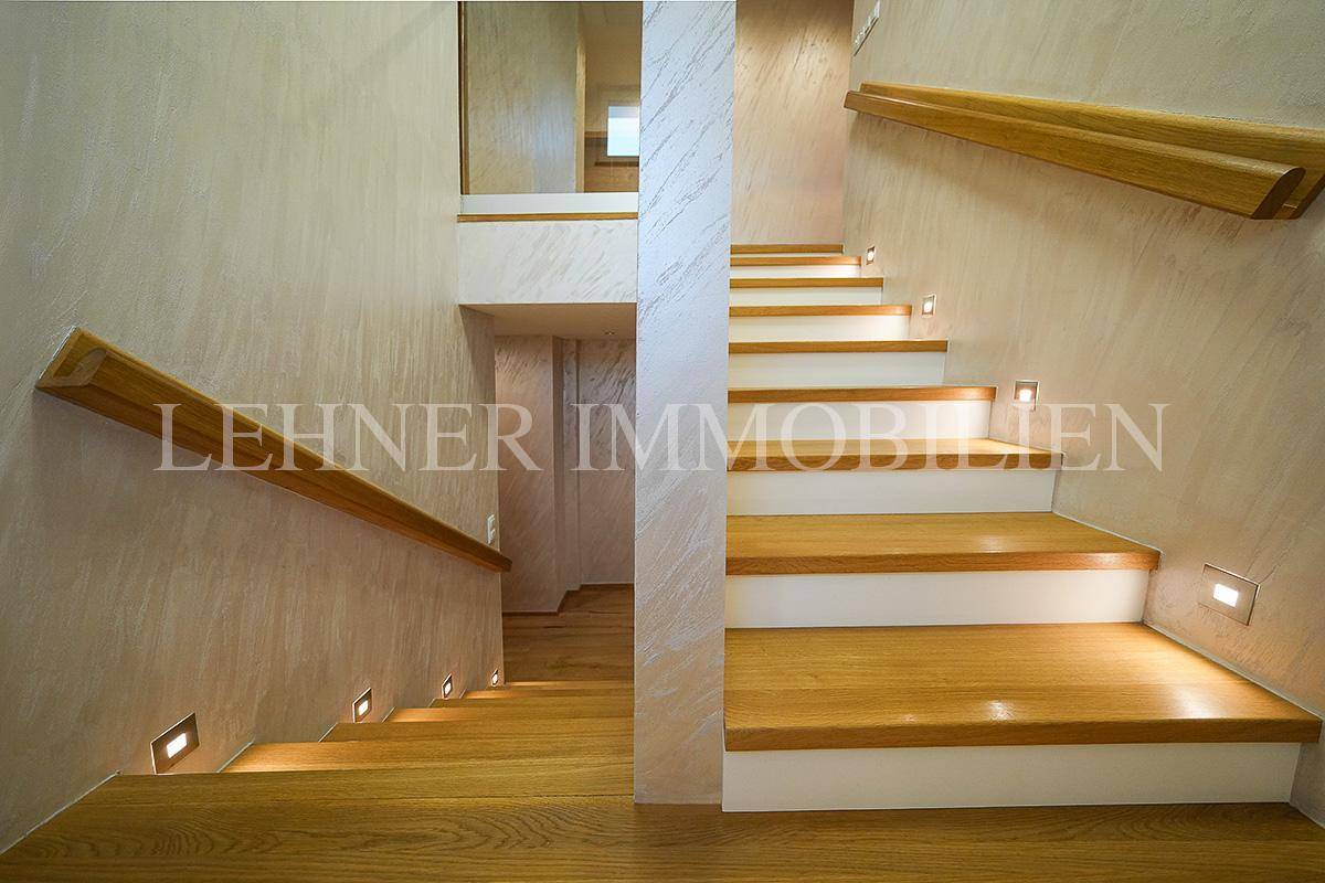 Moderner Treppenaufgang mit Holzstufen, Glasgeländer und integrierter Wandbeleuchtung.