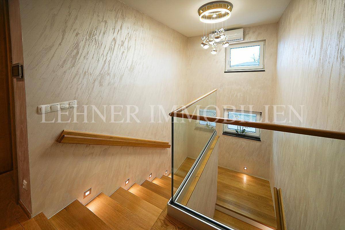 Treppenhaus mit Holzstufen, Glasgeländer und einer modernen Deckenleuchte, die für Helligkeit sorgt.