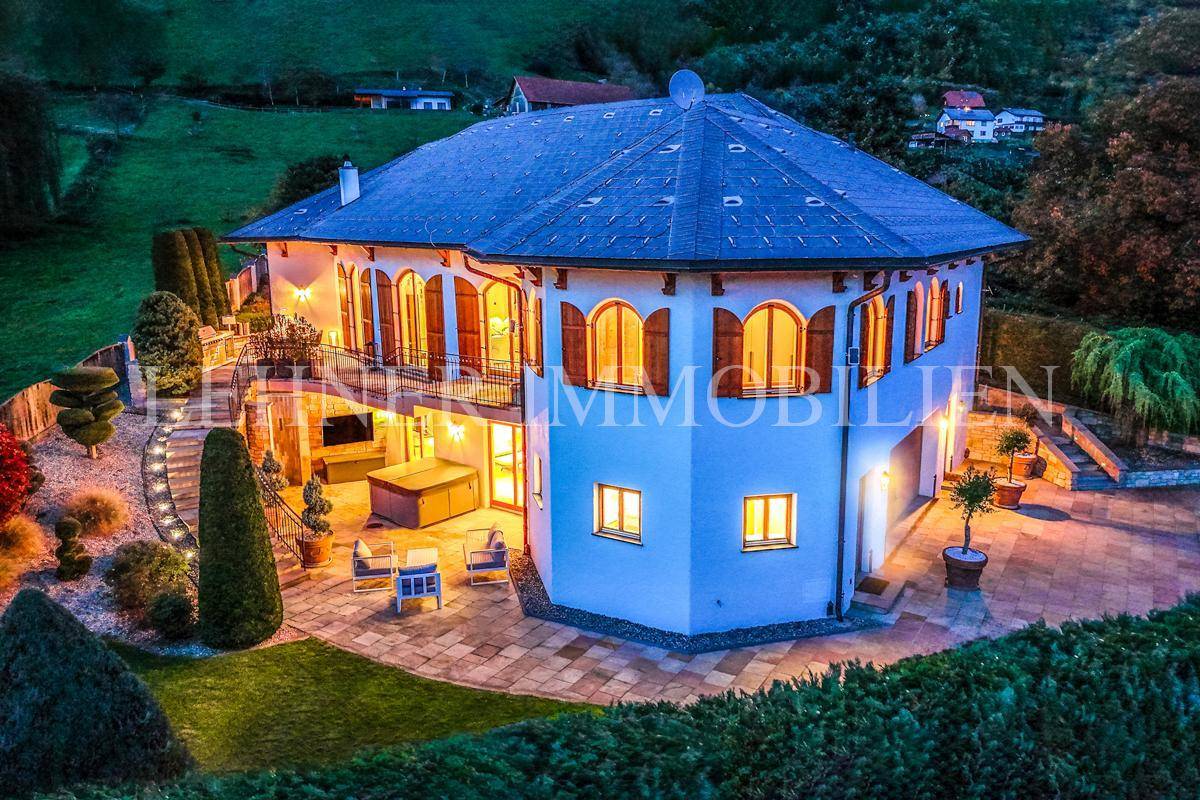 Beeindruckende Villa mit beleuchteter Terrasse und gepflegtem Garten, eingebettet in eine grüne Hügellandschaft.