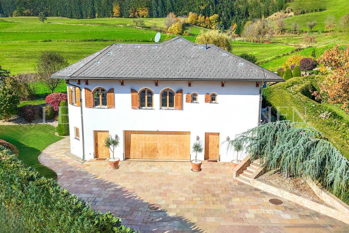 Rückansicht der Villa mit großem Garagentor und gepflastertem Vorplatz, umgeben von grüner Natur.