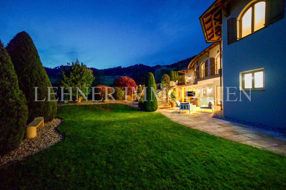 Gepflegter Rasenbereich mit angrenzender Terrasse und Blick auf die beleuchtete Villa bei Dämmerung.