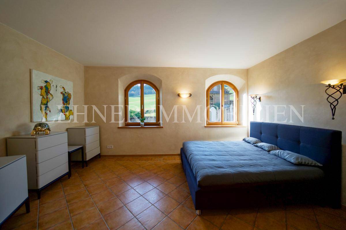 Helles Schlafzimmer mit blauem Bett, Kommoden und zwei Bogenfenstern mit Blick ins Grüne.