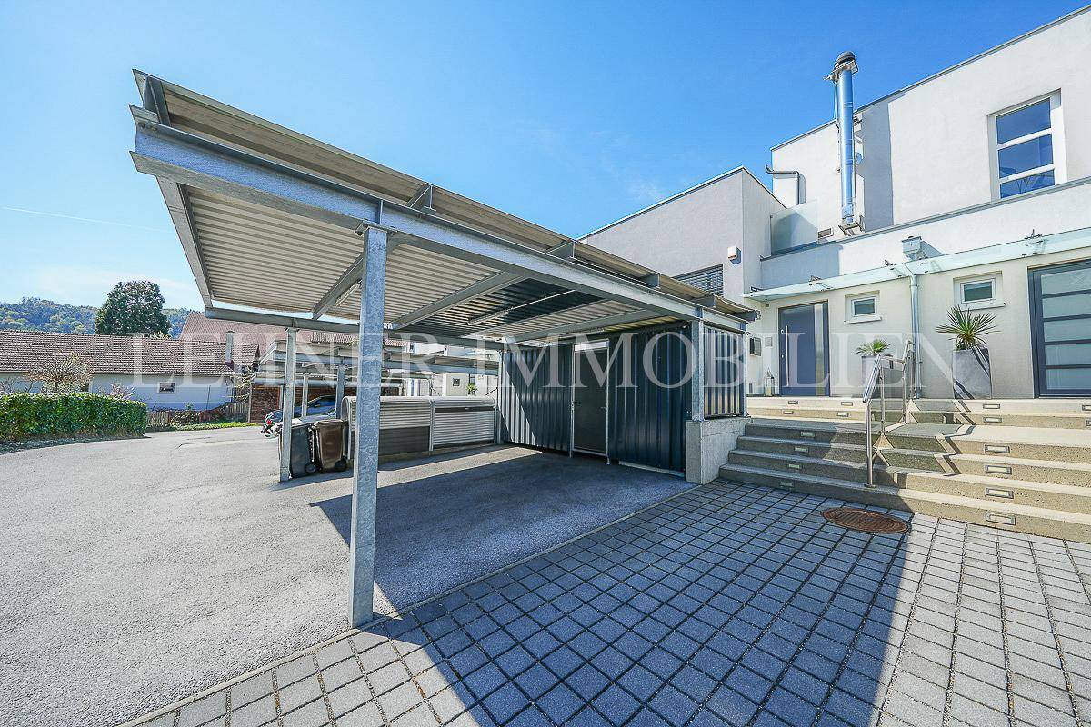 Moderner Carport vor einem Wohngebäude mit Außentreppe und gepflastertem Vorplatz.