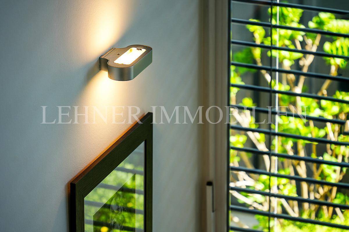 Detailaufnahme einer modernen Wandleuchte neben einem Fenster mit Jalousien und grüner Aussicht.