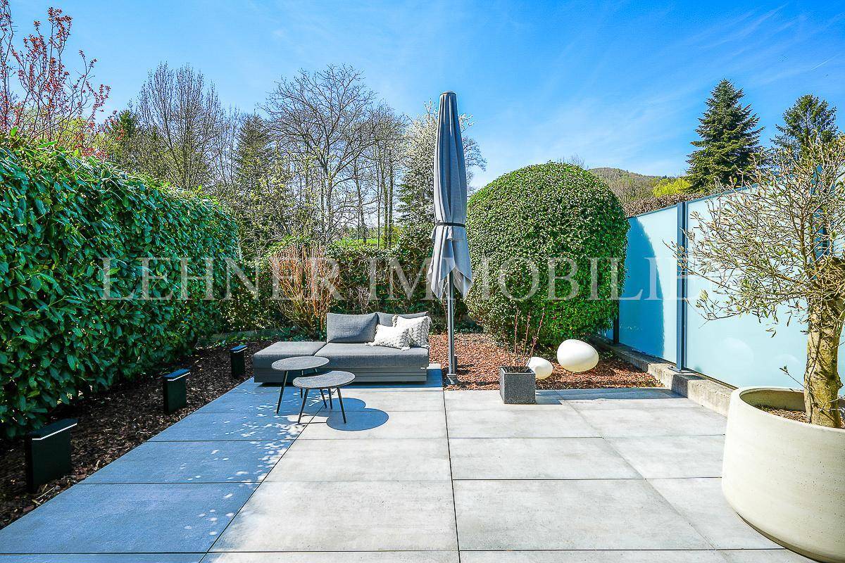 Sonniger Gartenbereich mit gepflasterter Fläche, Lounge-Möbeln und hoher Hecke für Privatsphäre.