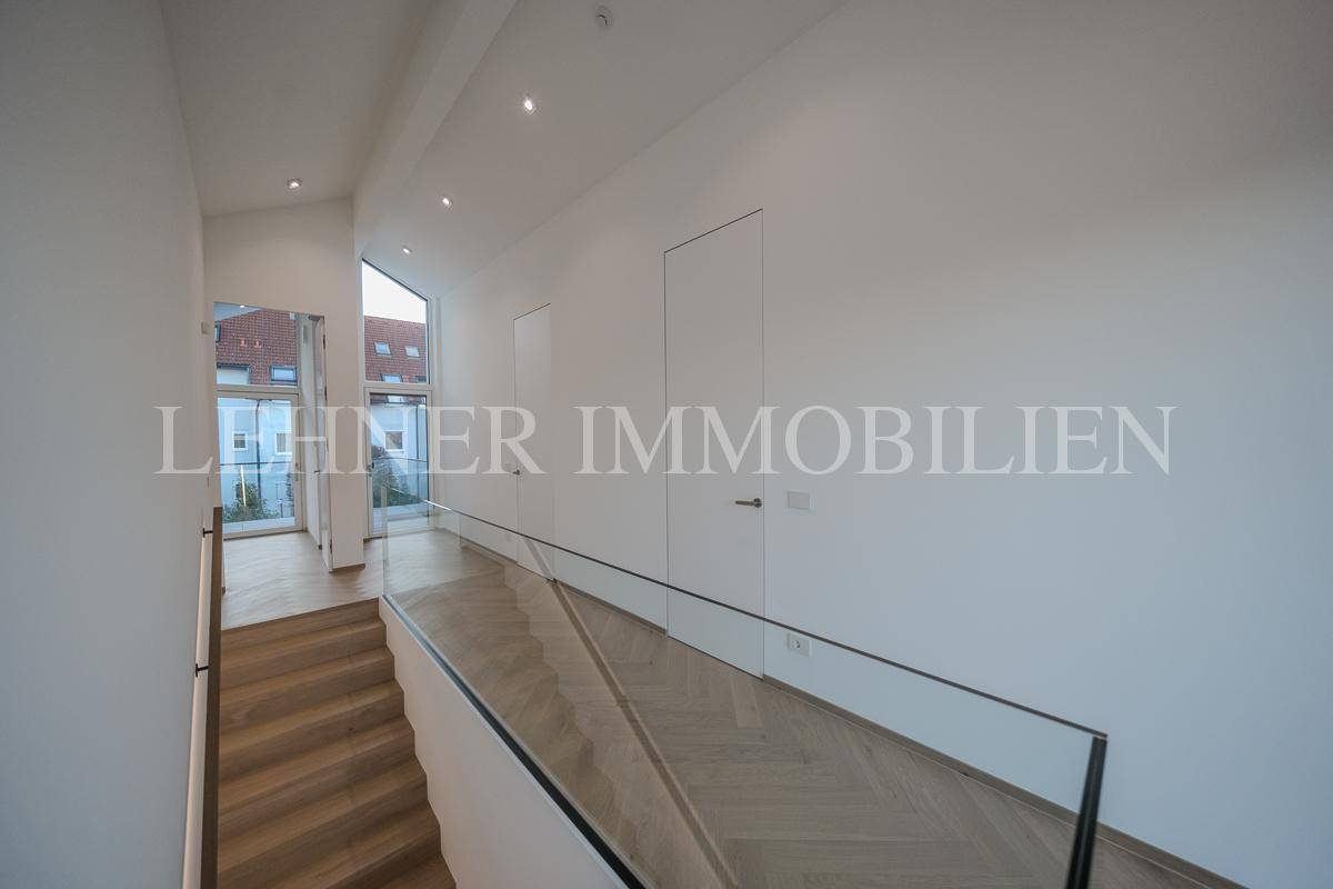 Moderner Flur mit Holztreppe, Glasgeländer und viel Tageslicht durch große Fenster.