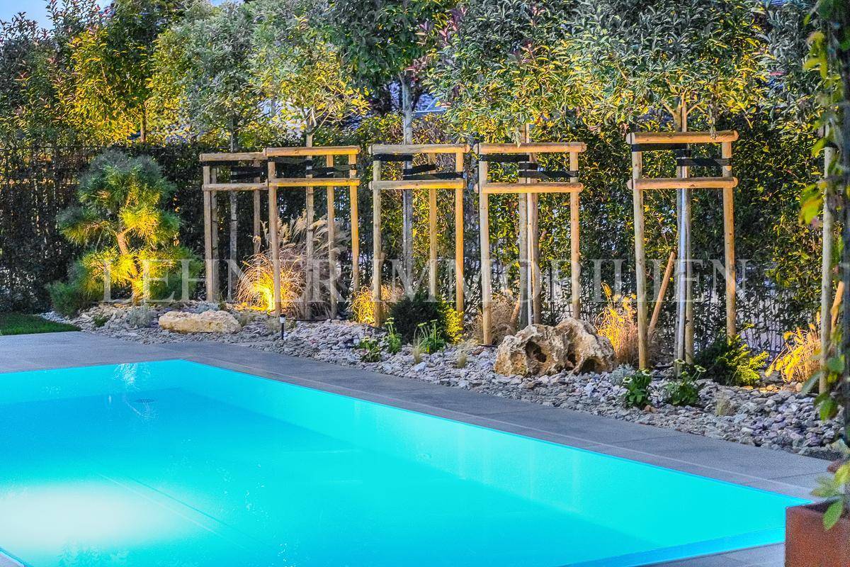 Stimmungsvoll beleuchteter Gartenbereich mit Zierpflanzen und dem Rand des Swimmingpools bei Nacht.