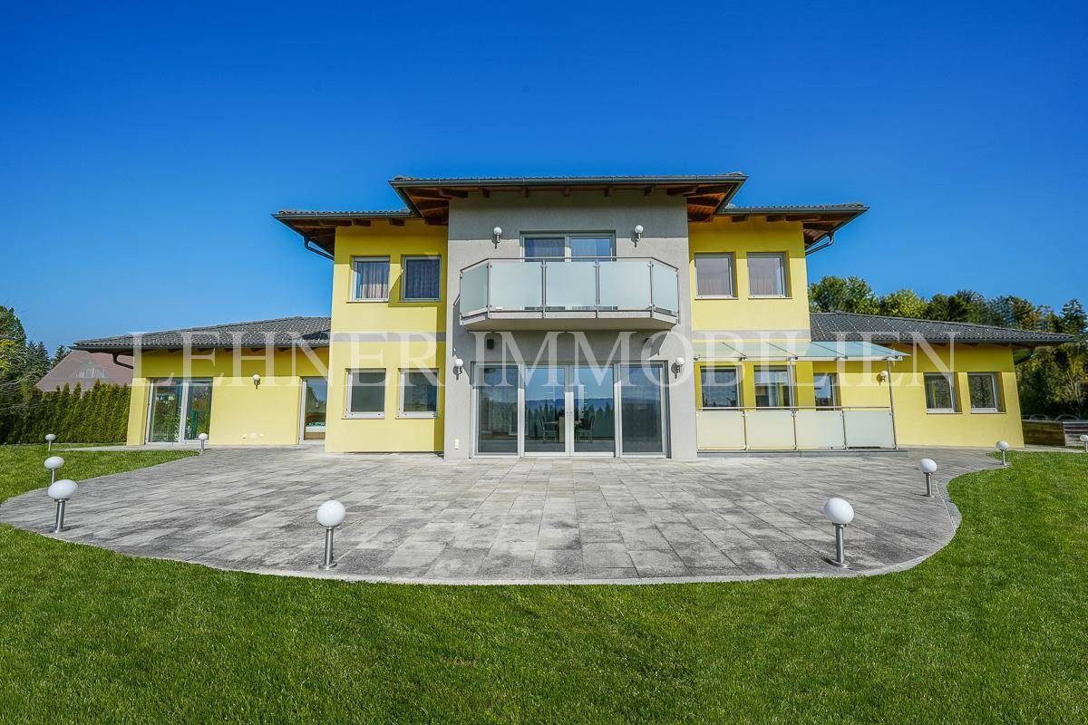 Frontansicht eines modernen Hauses mit gelber Fassade, gepflasterter Terrasse und Balkon.