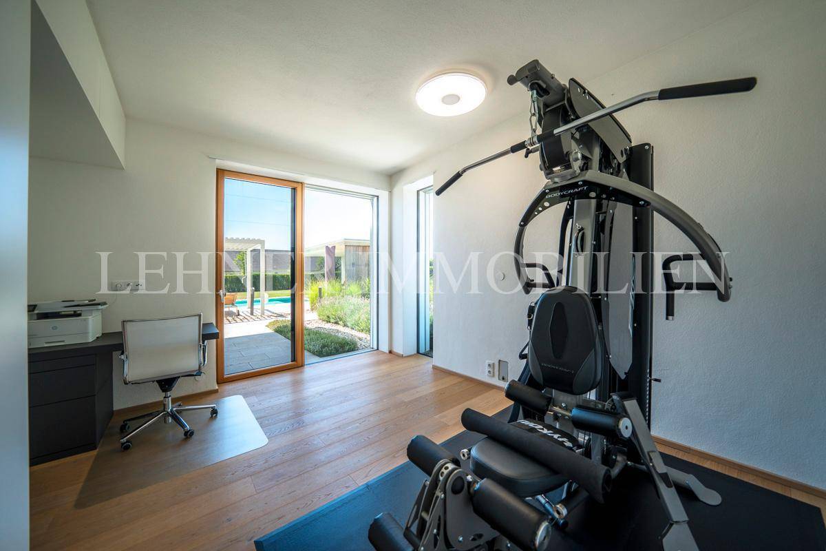 Heller Fitnessraum mit Trainingsgeräten, Schreibtisch und Blick durch die großen Fenster ins Grüne.