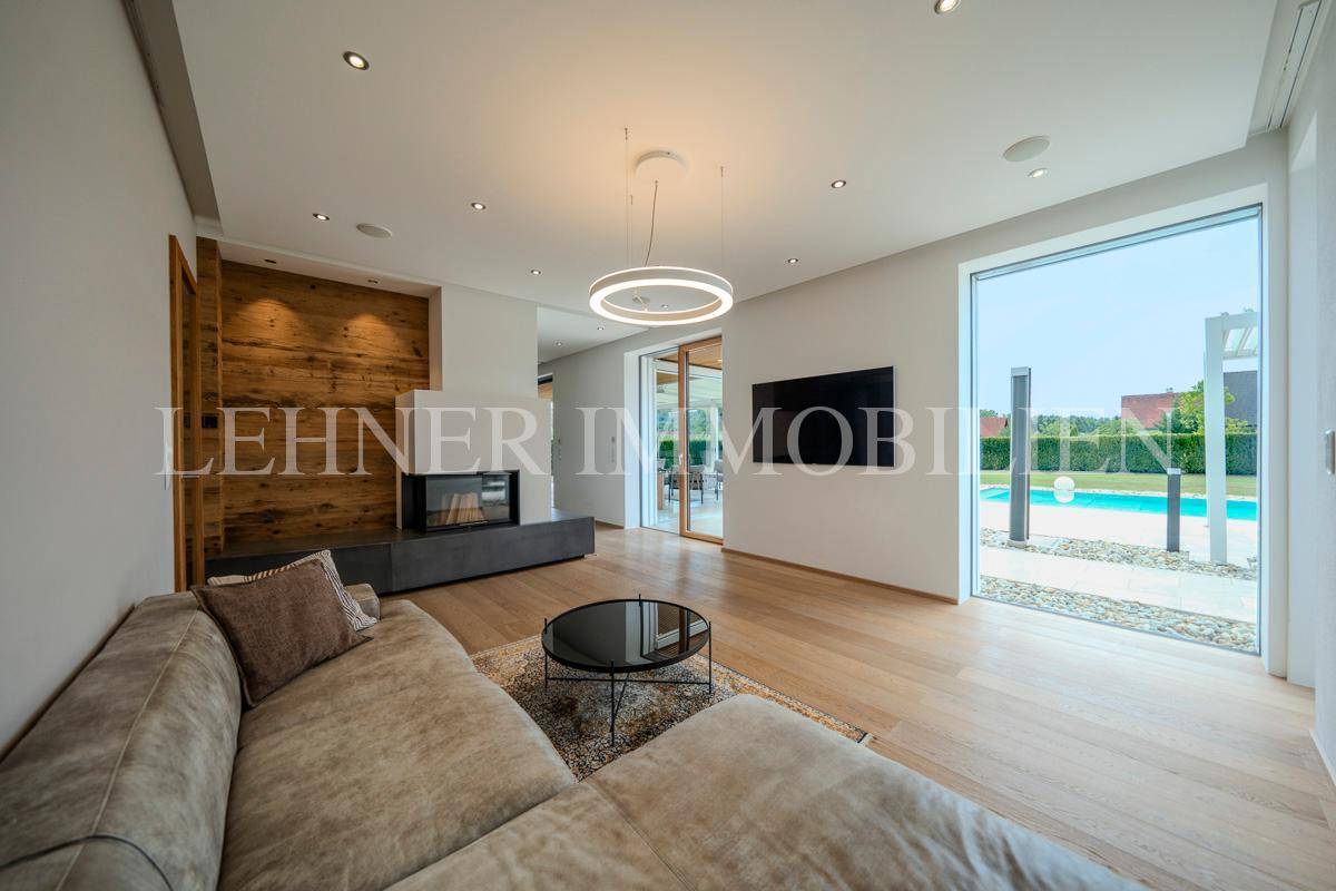 Helles Wohnzimmer mit großem Sofa, Kamin und Blick durch die Glasfront auf den Poolbereich.