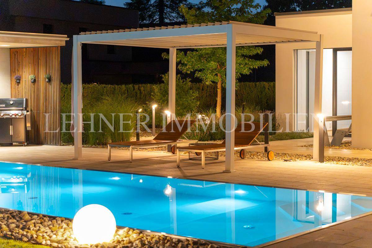 Gemütlicher Loungebereich unter einer Pergola neben dem beleuchteten Pool in der Abenddämmerung.