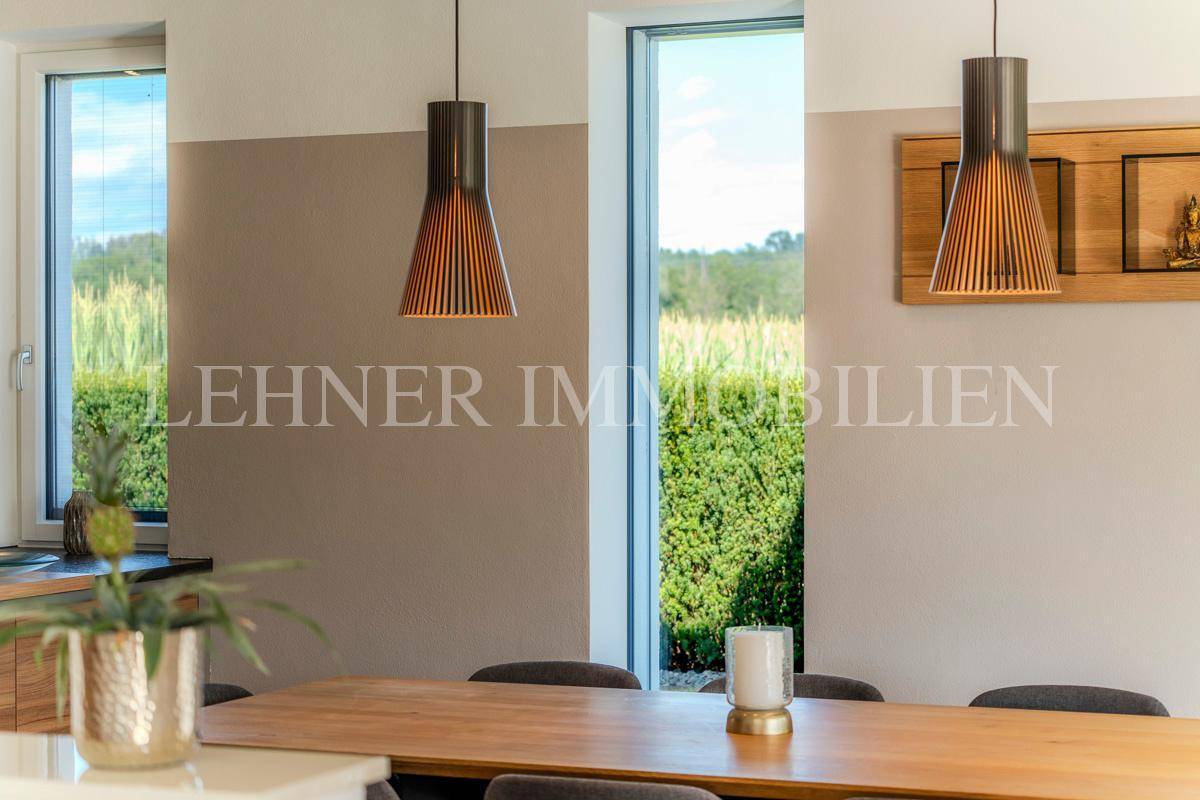 Essbereich mit Holztisch und Stühlen, umgeben von großen Fenstern mit Blick ins Grüne.