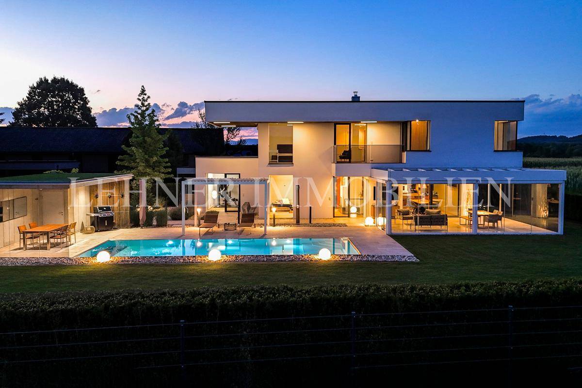 Moderne Villa mit beleuchtetem Pool, Garten und gemütlichem Essbereich im Freien bei Dämmerung.