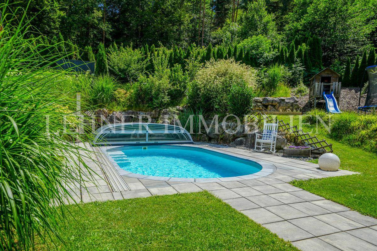 Ovaler Pool mit Abdeckung, umgeben von einer gepflasterten Fläche und grünem Garten.