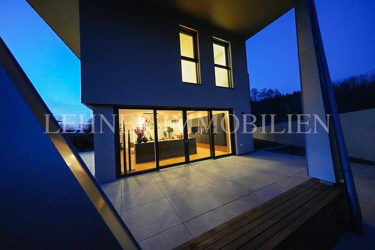 Beleuchtete Terrasse und Hausfassade bei Nacht, mit Blick auf die hellen Innenräume durch große Fenster.