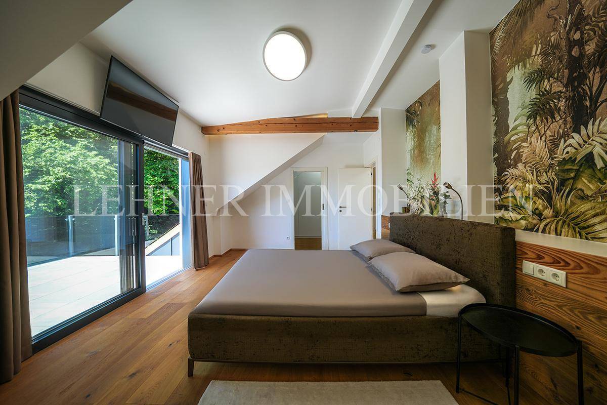 Helles Schlafzimmer mit Holzboden, freiliegenden Holzbalken und direktem Zugang zur Terrasse mit grüner Aussicht.
