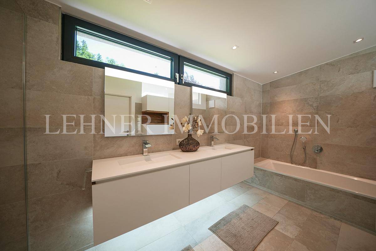 Modernes Badezimmer mit großen grauen Fliesen, Doppelwaschbecken und Badewanne unter einem breiten Fenster.