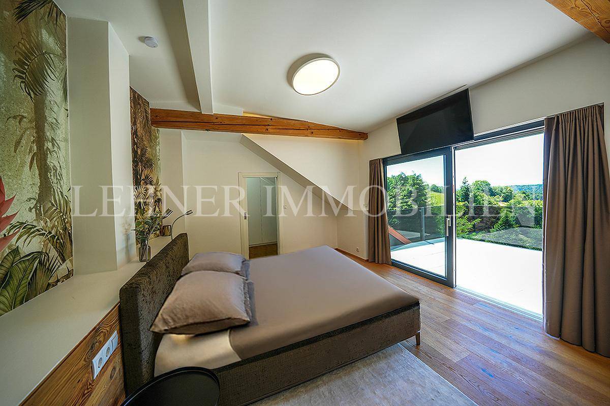 Helles Schlafzimmer mit Holzboden, freiliegenden Holzbalken und direktem Zugang zur Terrasse mit grüner Aussicht.
