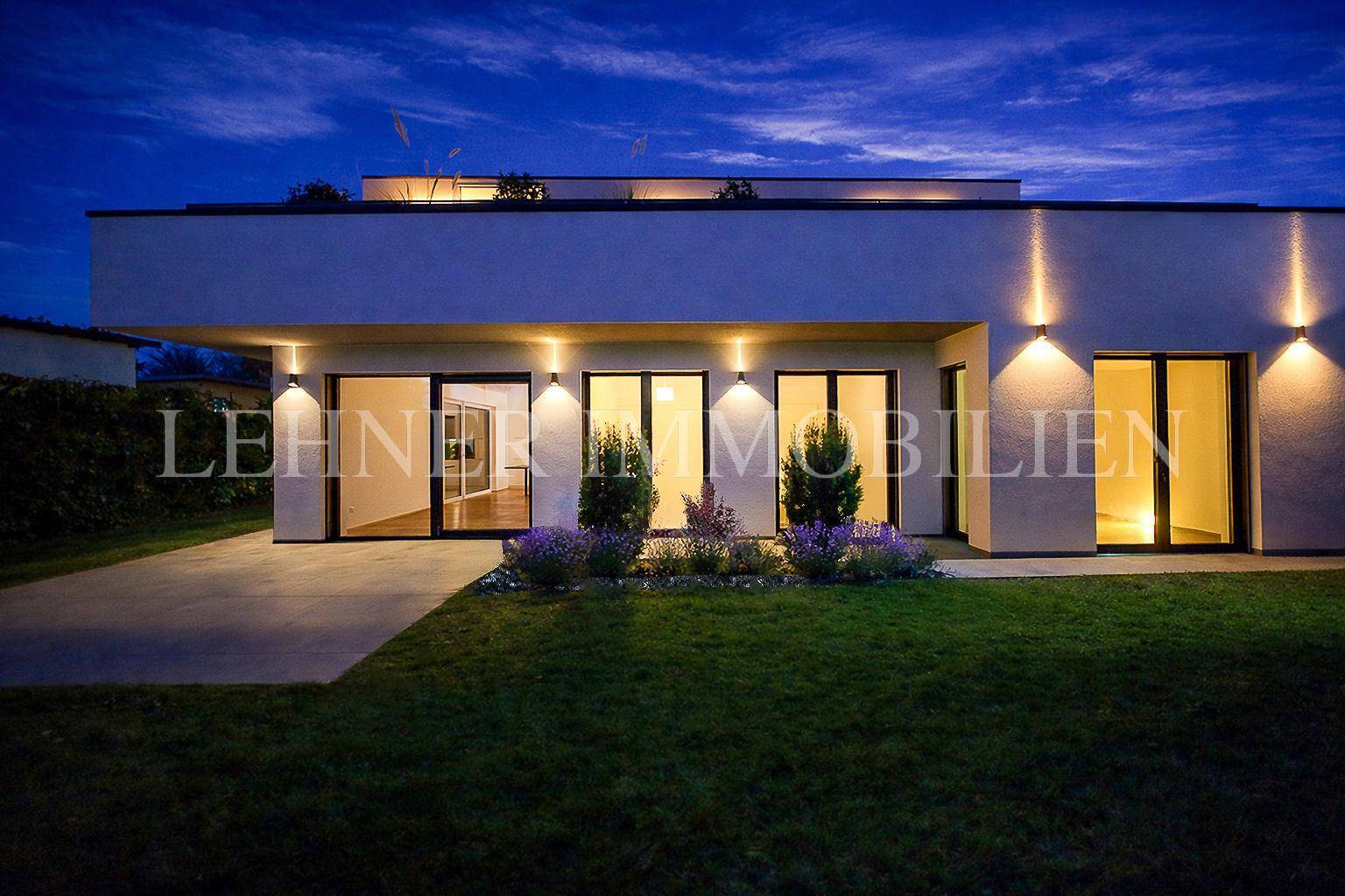 Modernes Haus bei Nacht mit beleuchteter Fassade und kleinen Pflanzenbeeten im Garten.