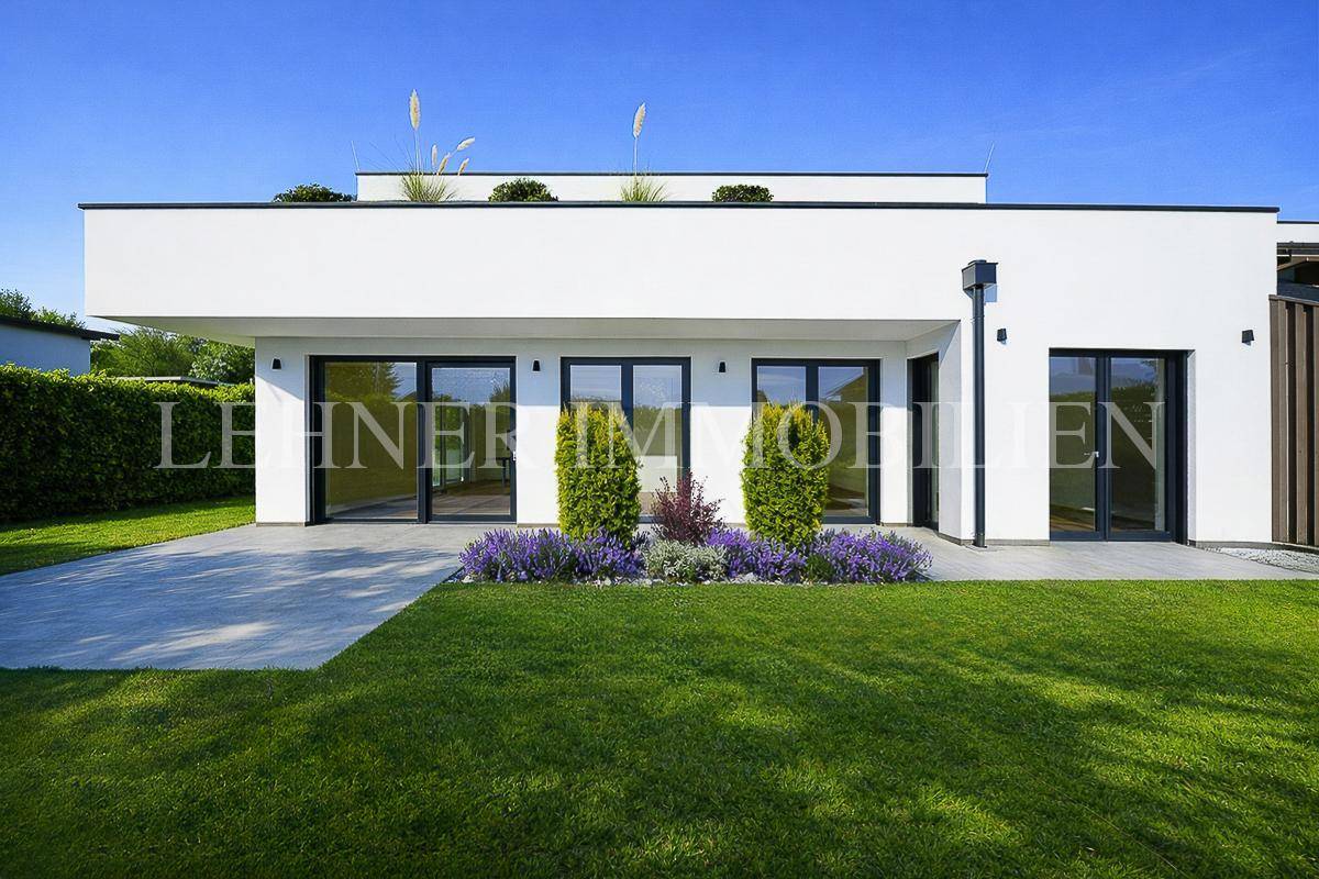 Moderne Hausfassade mit großen Fenstern, gepflegtem Rasen und blühenden Pflanzenbeeten.