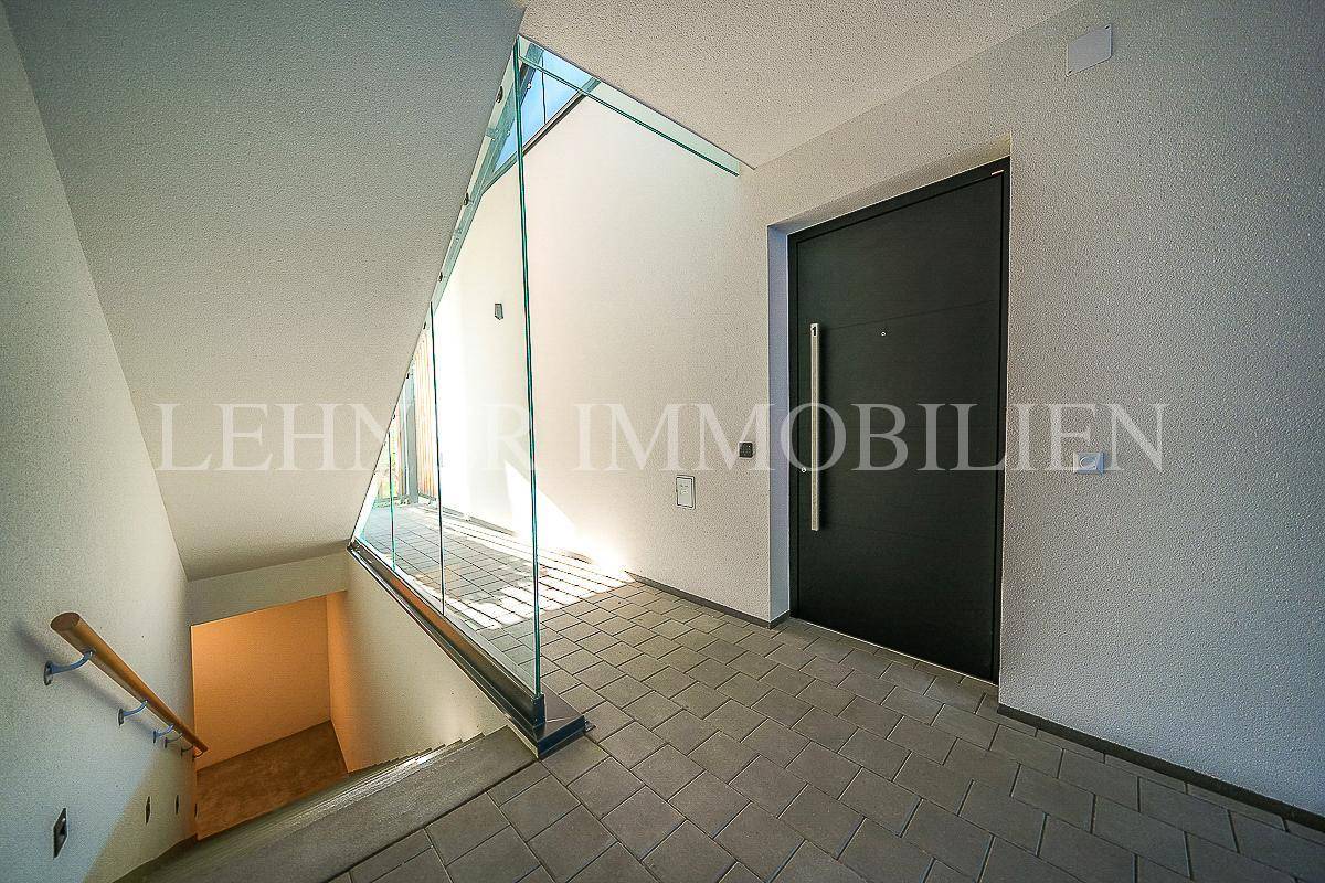 Heller Eingangsbereich mit moderner Haustür, Fliesenboden und Treppe mit Glasgeländer zum Untergeschoss.