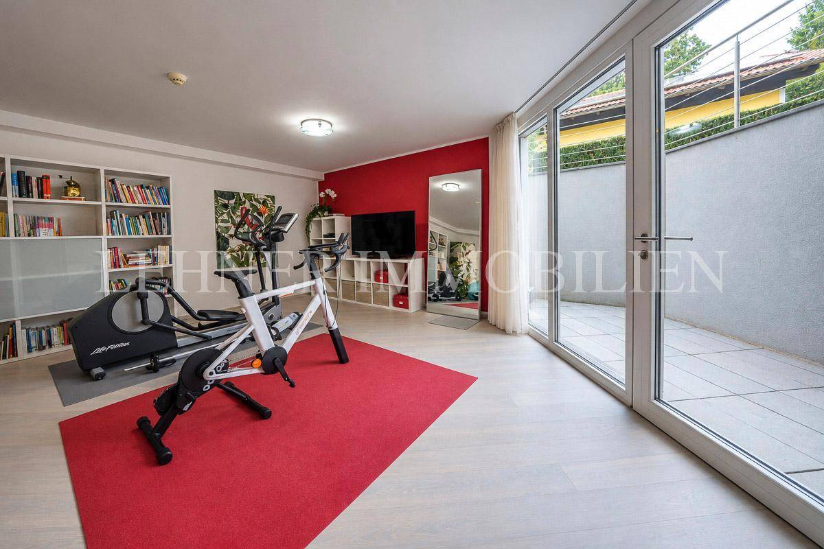 Heller Fitnessraum mit Sportgeräten, Bücherregal und direktem Zugang zur Terrasse.