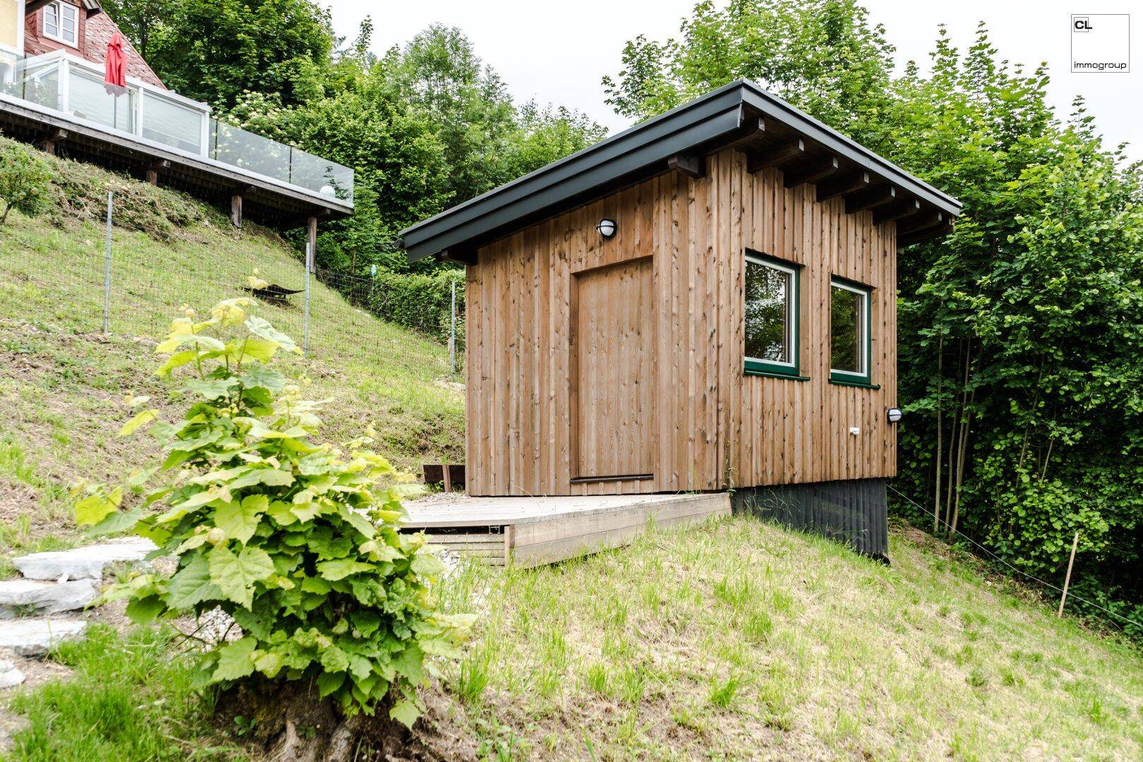 Kleines Holzhaus auf einem grünen Hügel, umgeben von Bäumen und Natur.