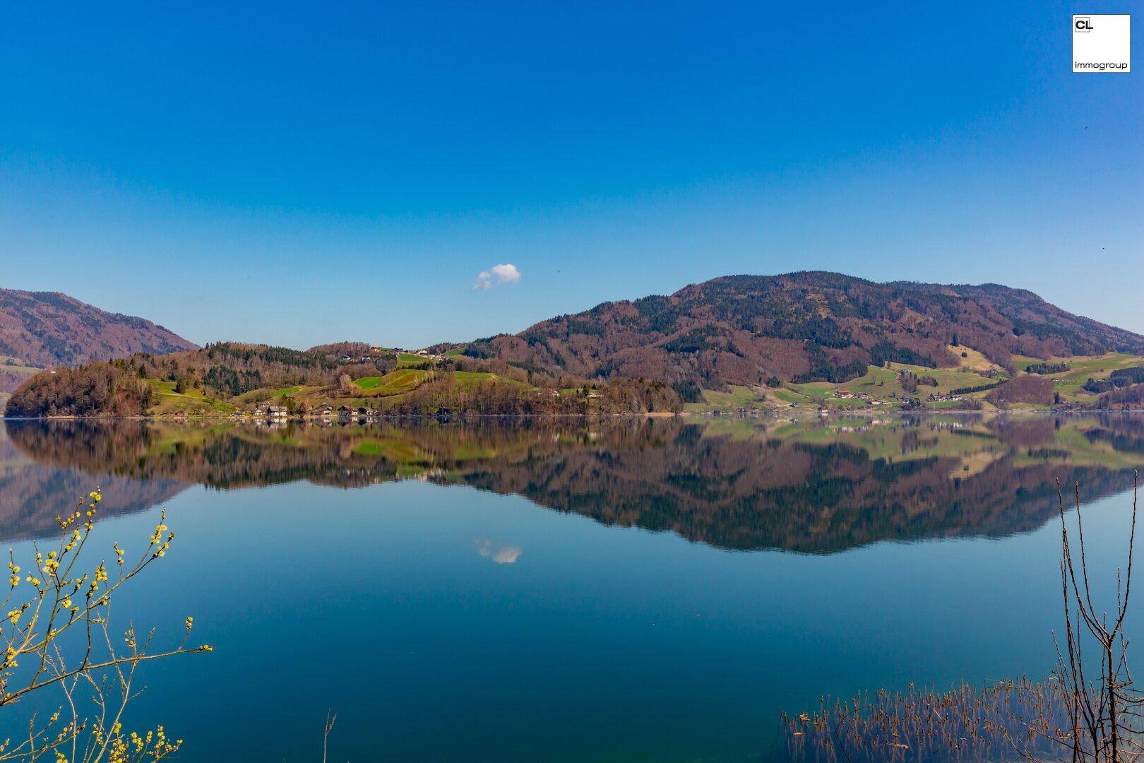 Ruhiger See mit spiegelnder Oberfläche und bewaldeten Hügeln unter klarem blauem Himmel.
