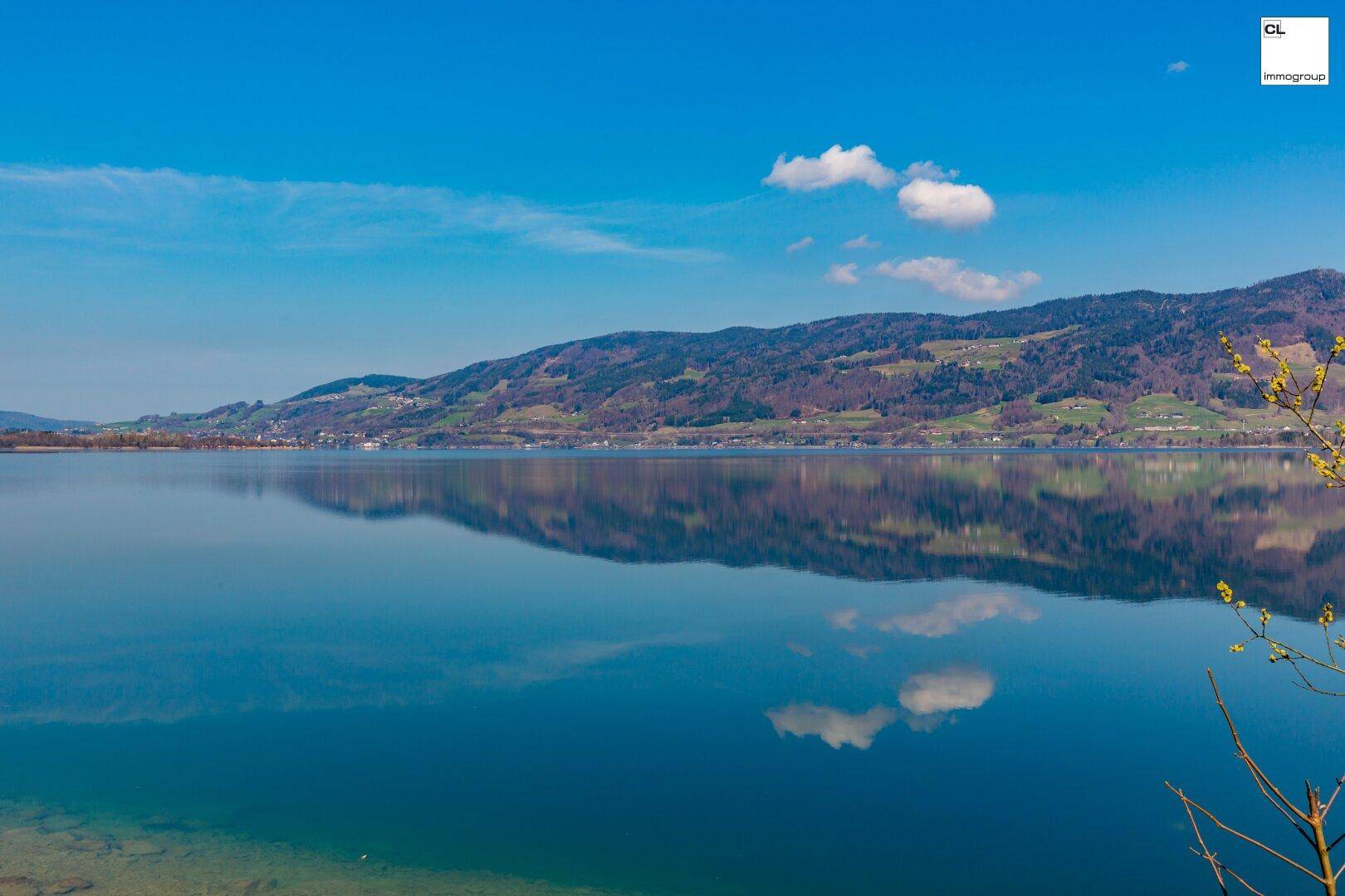 Breiter Blick über einen klaren See, umgeben von grünen Hügeln und einem strahlend blauen Himmel.
