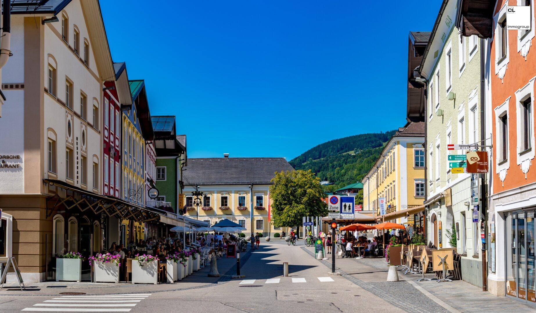 Belebte Straße in einer charmanten Stadt mit traditionellen Gebäuden und Außengastronomie unter blauem Himmel.