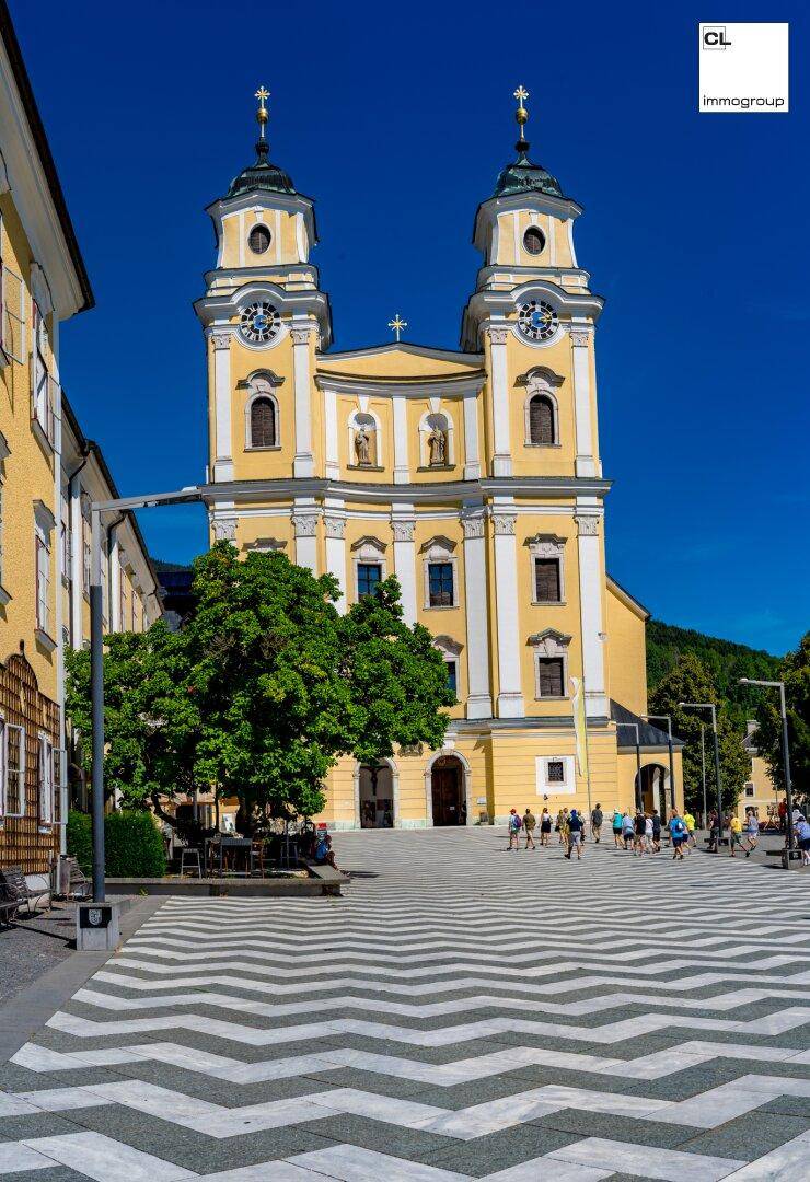 Imposante gelbe Kirche mit zwei Türmen und Uhren, umgeben von einem belebten Platz mit Menschen.