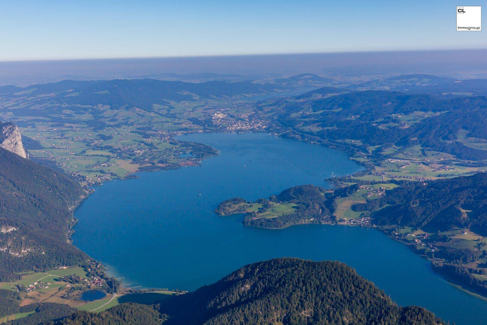 Weitläufige Luftaufnahme eines großen Sees, eingebettet in eine grüne Hügellandschaft mit vielen kleinen Ortschaften.