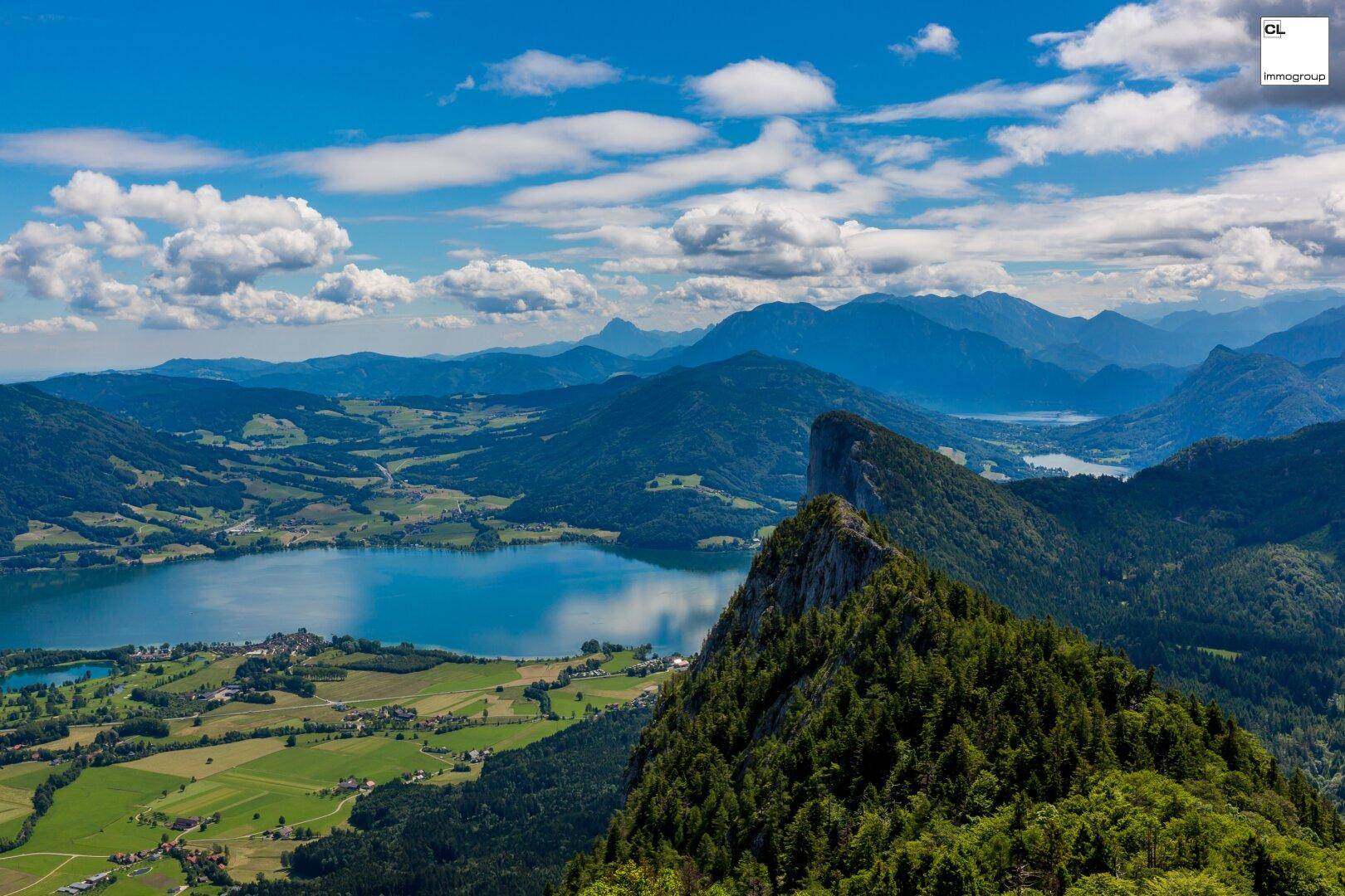 Spektakuläre Luftaufnahme einer Berglandschaft mit einem glitzernden See und weiten grünen Tälern unter Wolken.