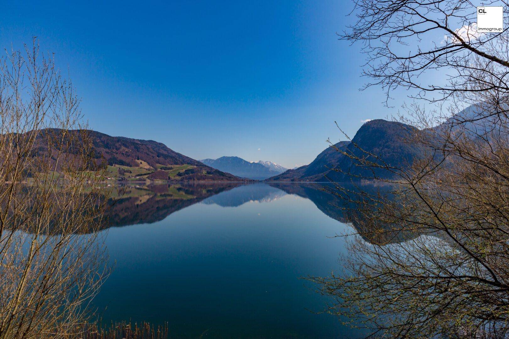 Panoramablick über einen stillen See, umrahmt von bewaldeten Hügeln und schneebedeckten Bergen in der Ferne.