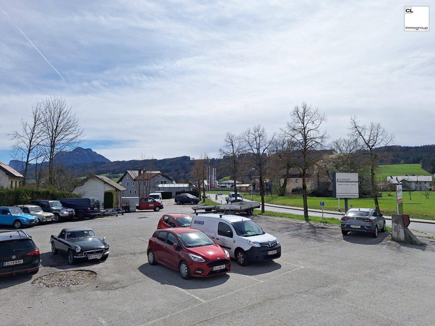 Großer Parkplatz mit verschiedenen Autos, umgeben von grüner Landschaft und Bergen im Hintergrund.