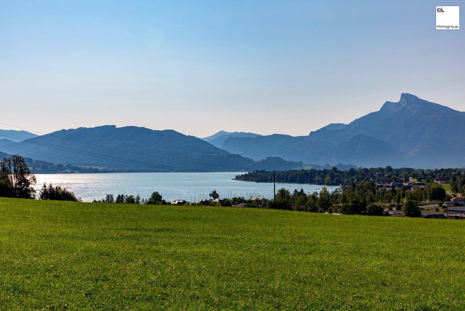 Weitläufiger Panoramablick über einen glitzernden See, umgeben von majestätischen Bergen und grünen Wiesen.