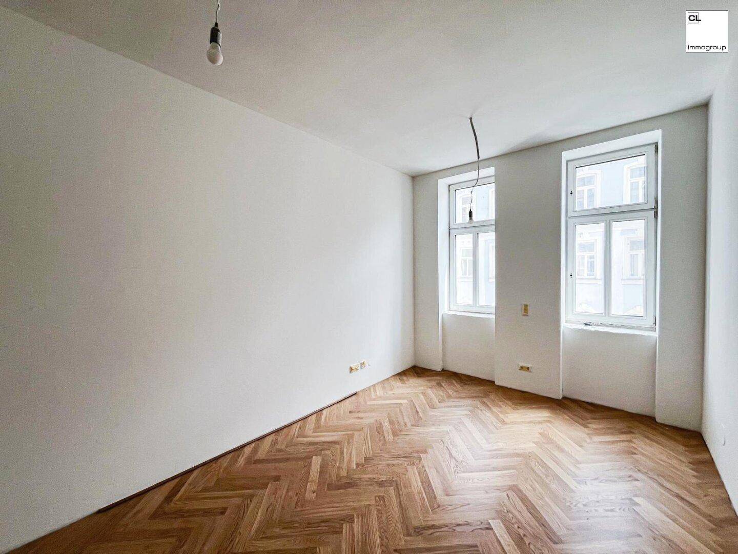 Lichtdurchflutetes Zimmer mit klassischem Fischgrätparkett und zwei großen Fenstern.