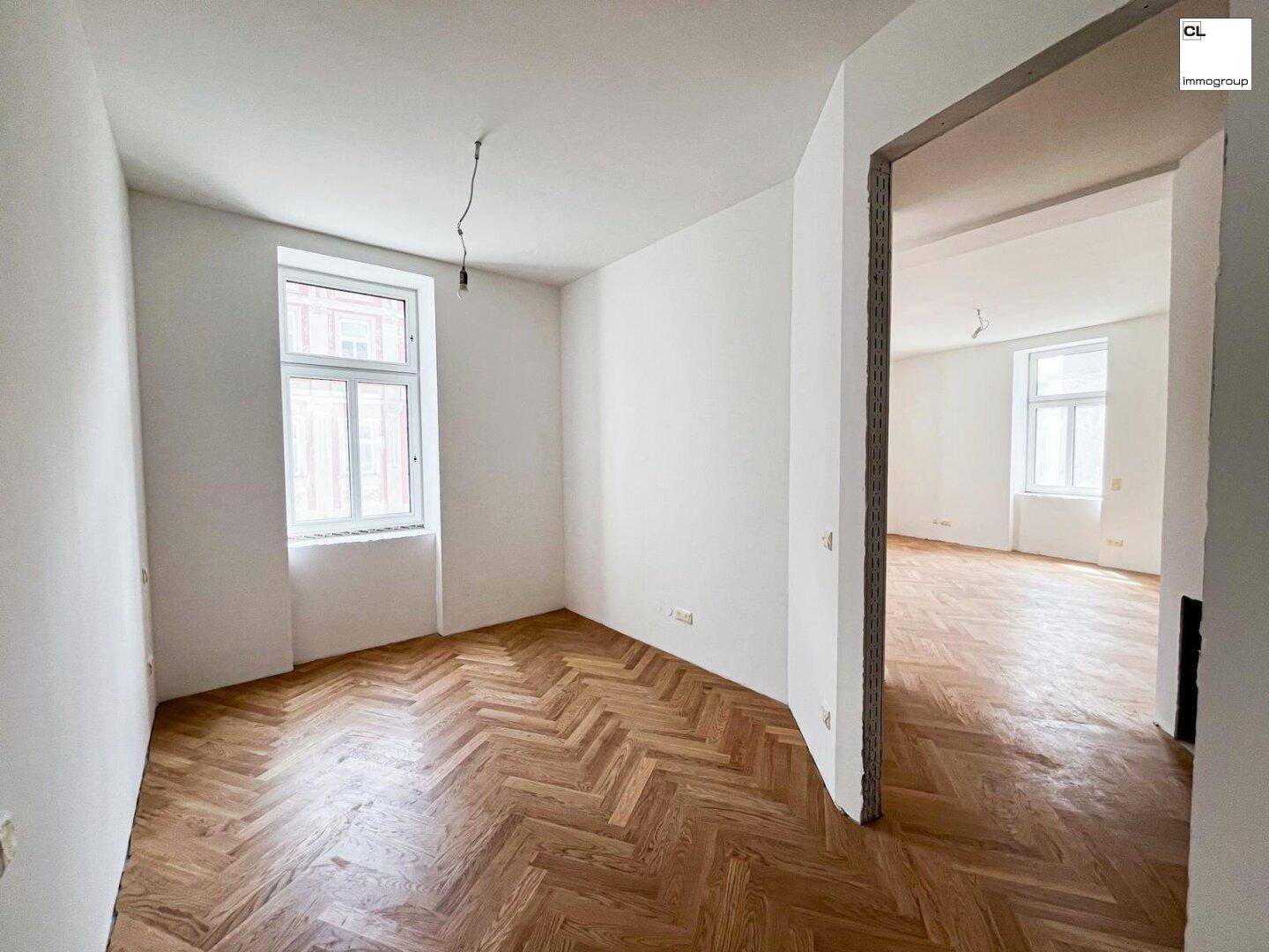 Geräumiges Zimmer mit Fischgrätparkett und offenem Zugang zu einem weiteren hellen Raum.