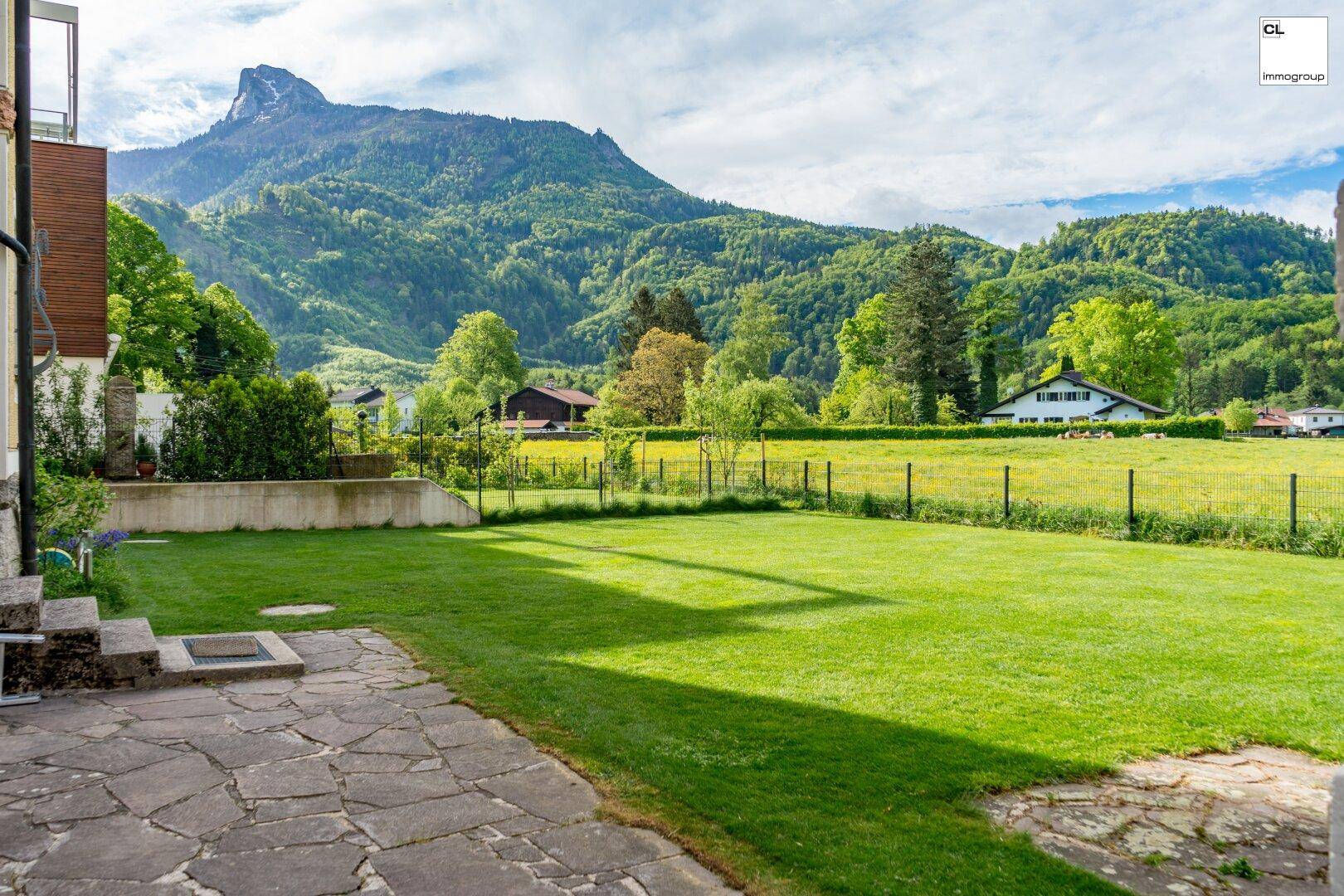 Weitläufiger Garten mit Rasenfläche und Steinterrasse, umgeben von grünen Bergen und Feldern.