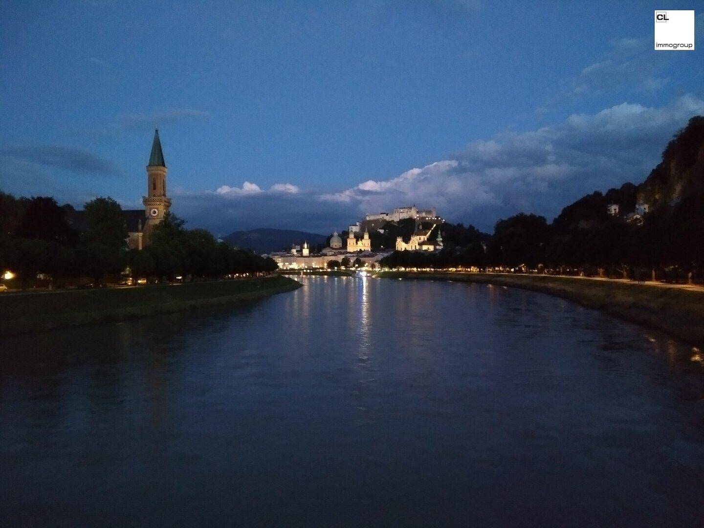 Panoramablick auf eine Stadt bei Nacht mit einem Fluss, einer Burg und beleuchteten Gebäuden.