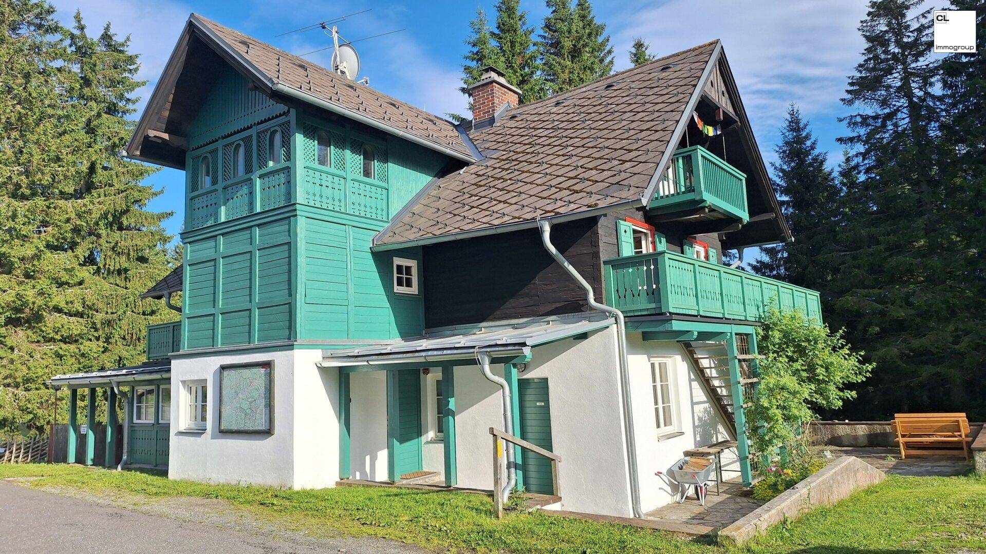 Charmantes Landhaus mit markanter grüner Holzverkleidung, traditionellem Satteldach und umgebenden Nadelbäumen.
