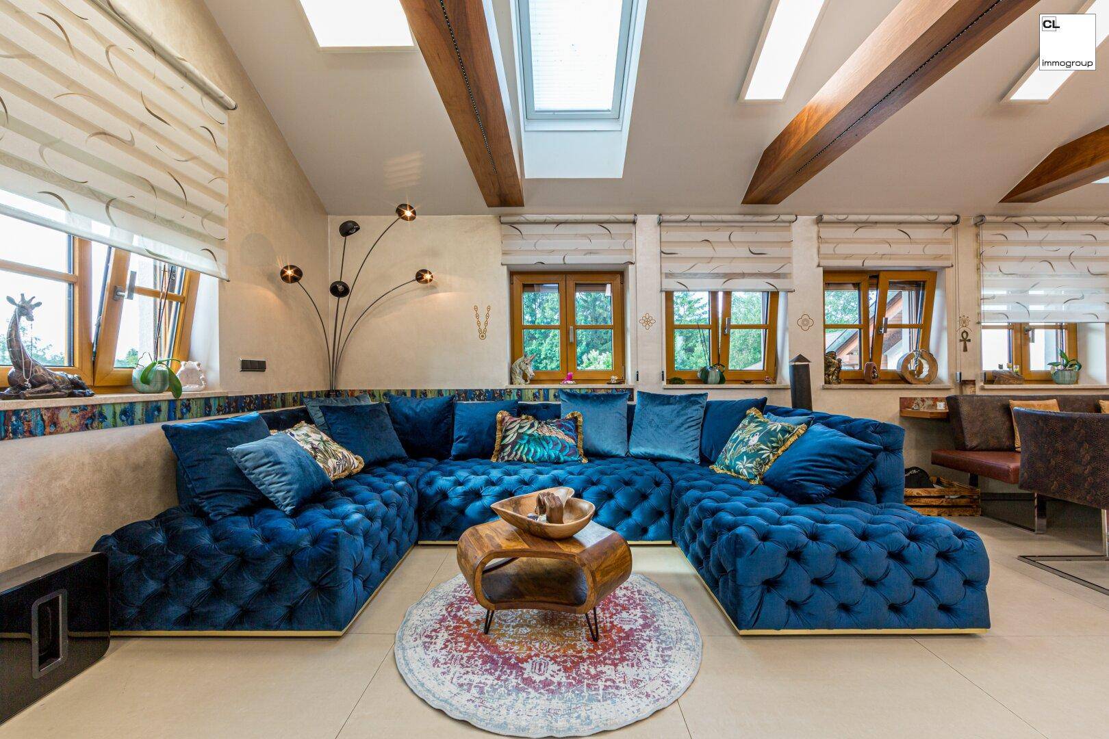 Gemütliches Wohnzimmer mit großem blauen Sofa, Holzbalken und Blick ins Grüne.