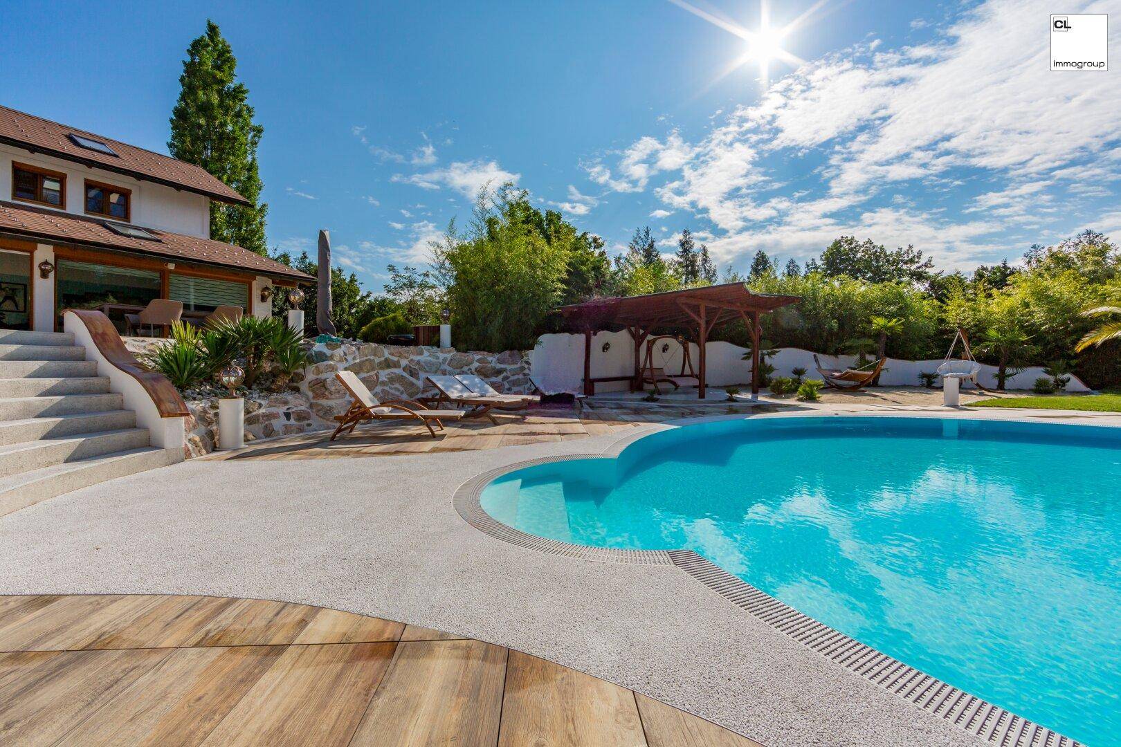 Großer Außenpool mit gepflasterter Terrasse und Liegestühlen unter blauem Himmel.