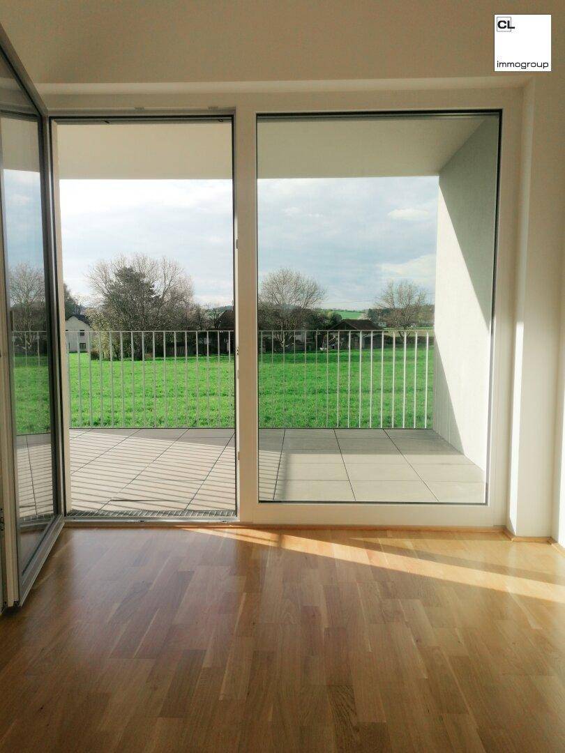 Blick aus einem hellen Zimmer auf eine Terrasse mit weitem Grünblick und viel Natur.