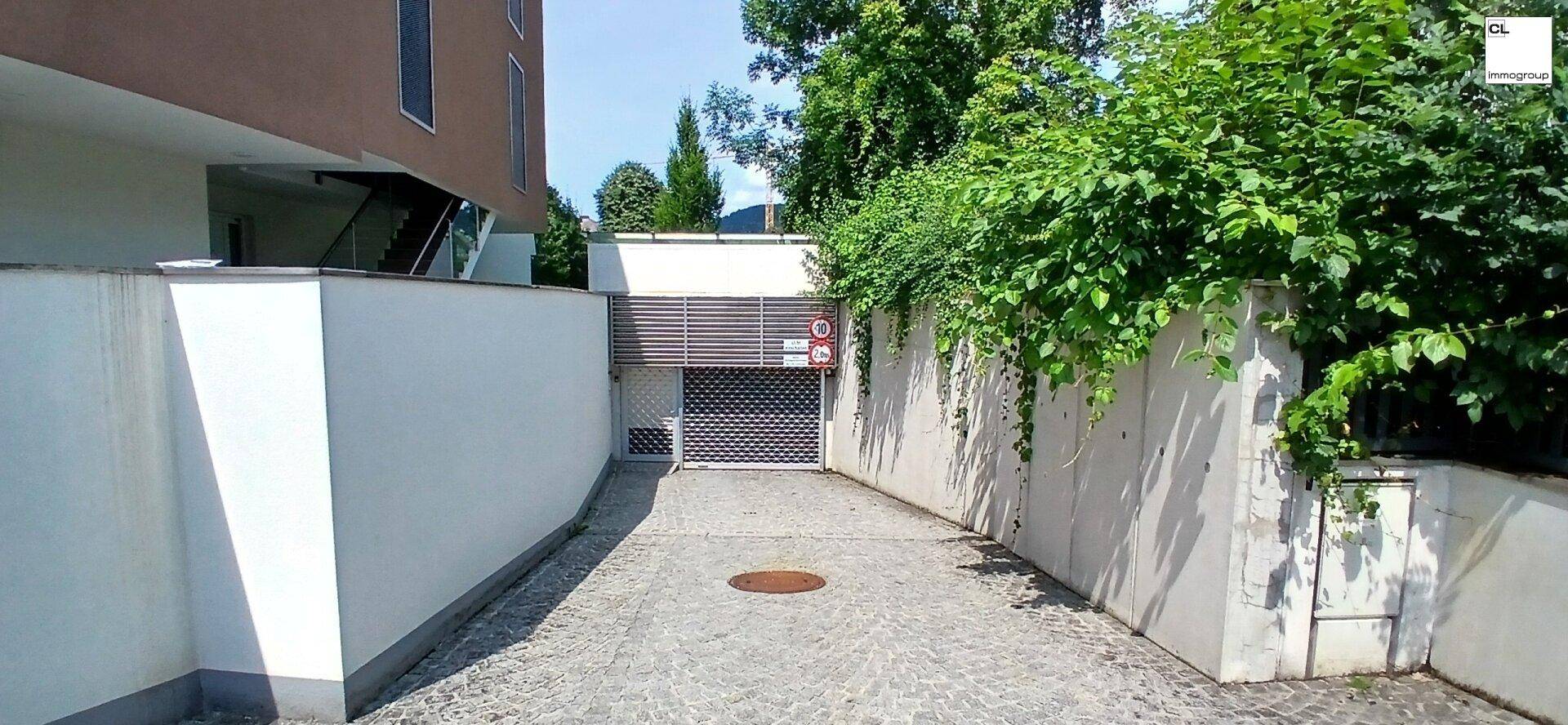 Zufahrt zur Tiefgarage, umgeben von weißen Mauern und üppigem Grün, mit Kopfsteinpflaster.