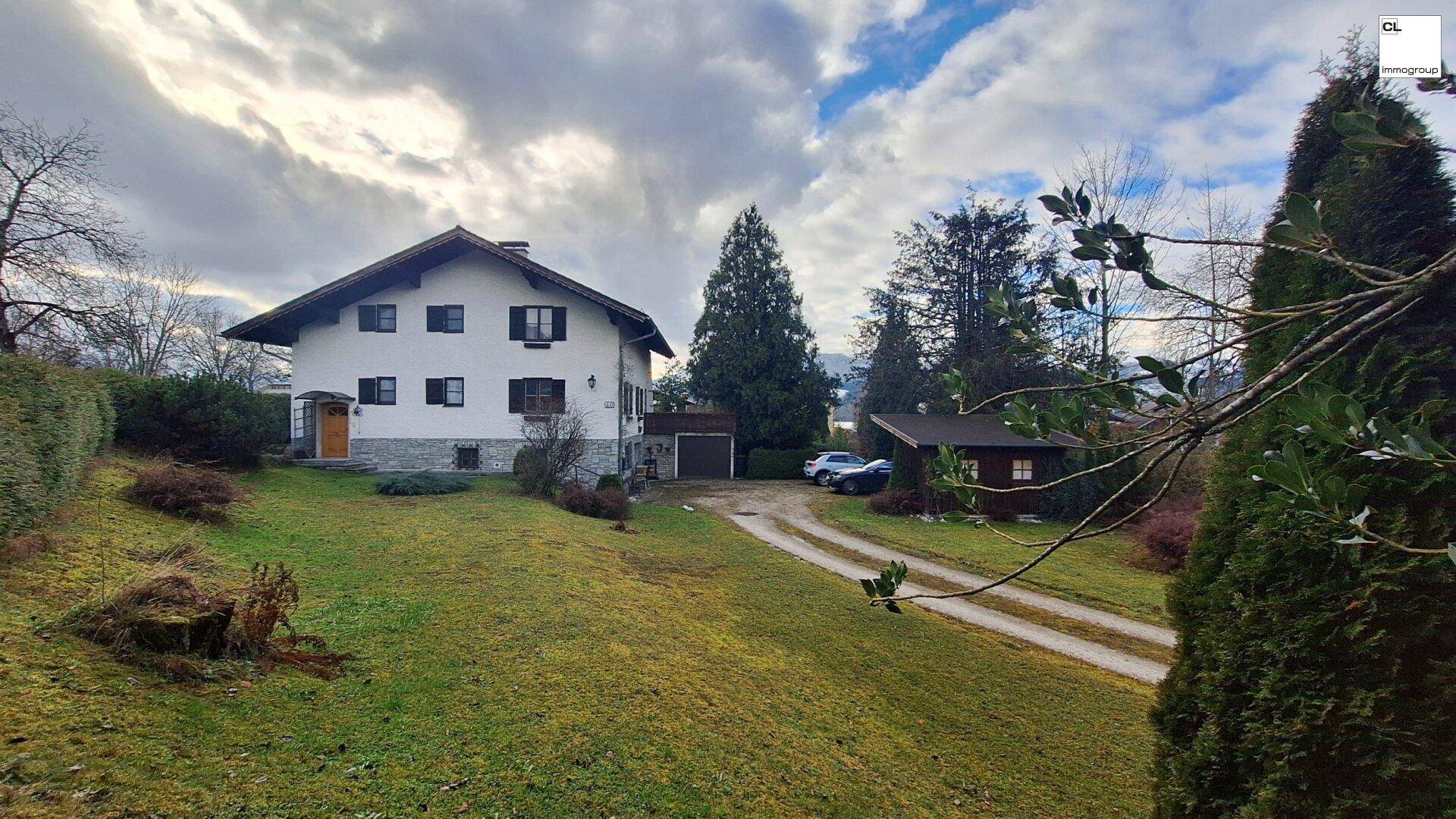 Charmantes Haus mit großem Vorgarten, langer Auffahrt und umgebenden Bäumen unter einem bewölkten Himmel.
