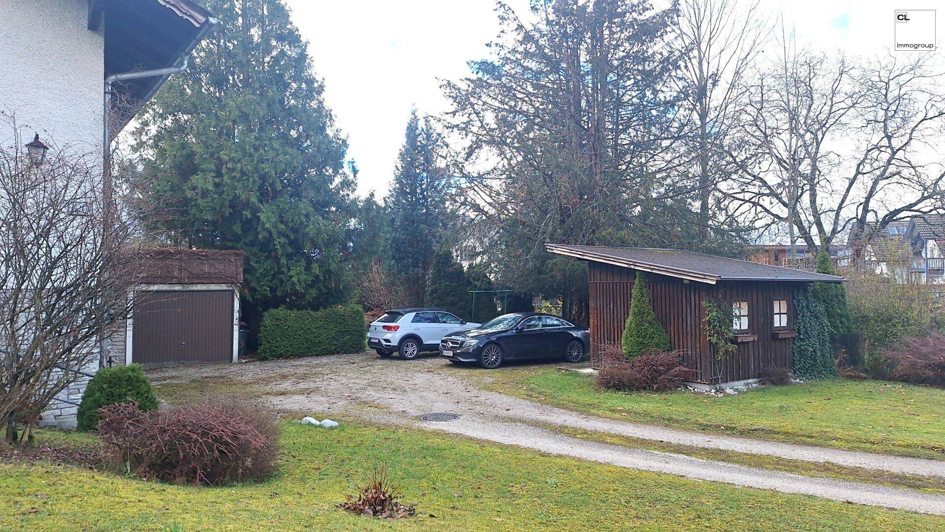 Parkplatz mit zwei Autos und einem kleinen Holzschuppen neben dem Haus, umgeben von Bäumen.